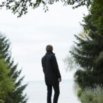Man in a coat standing on path, surrounded by trees, overlooking a serene lake on a cloudy day.