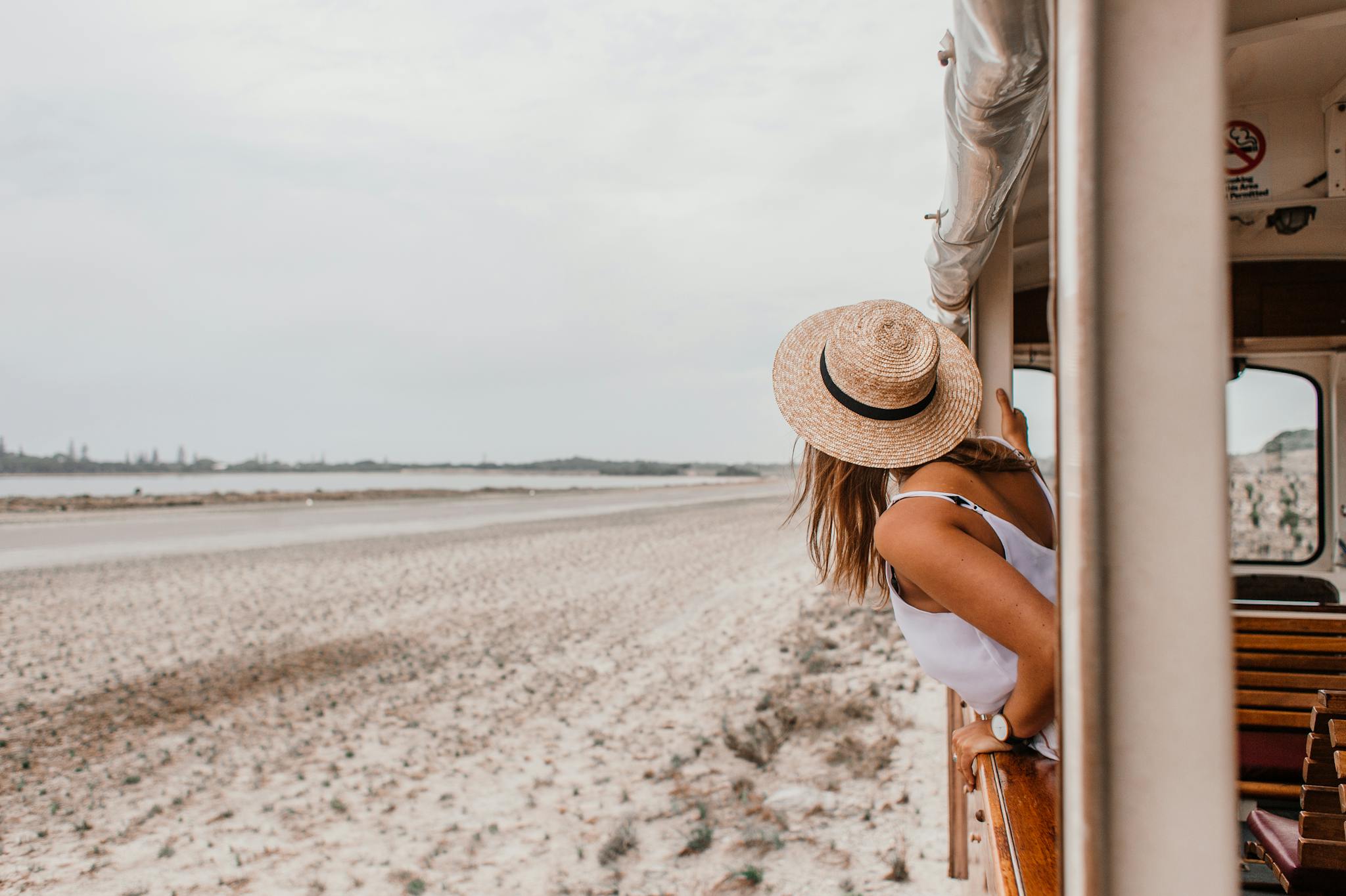 Woman in straw hat leans out of a train window, enjoying a beachside journey.