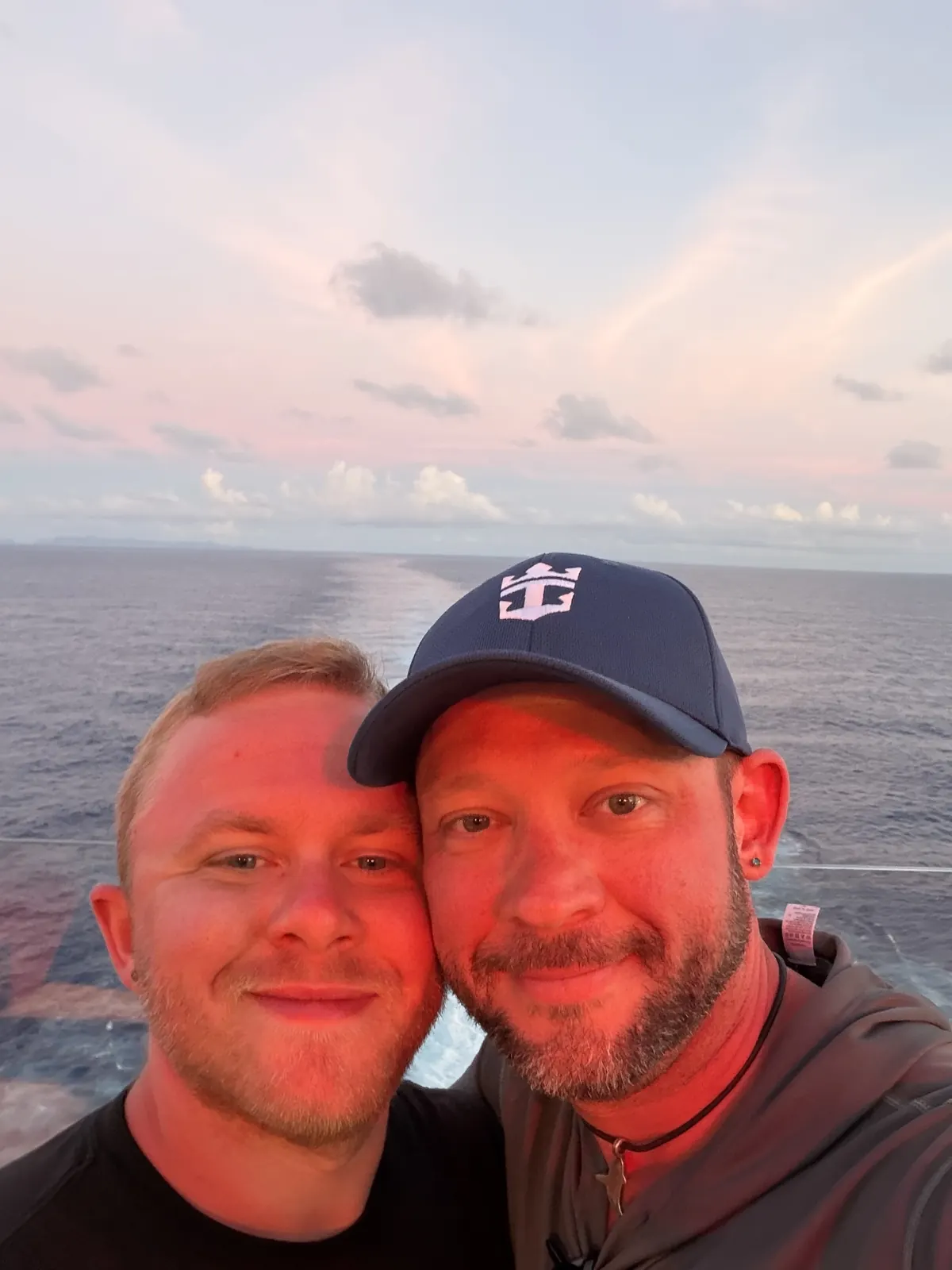 Fox and his husband Brandon taking a selfie on a cruise ship deck at sunset, Fox wearing a Royal Caribbean cap